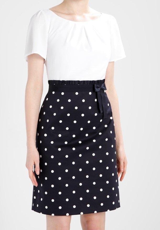 Femme portant une jupe bleu marine longueur genou à pois blancs avec un petit nœud, associée à un chemisier blanc à manches courtes.