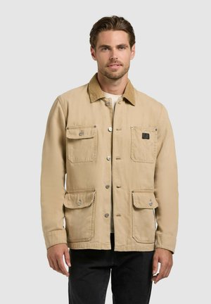 Beige színű, négy első zsebbel rendelkező, gombos dzsekit viselő férfi, fekete nadrágban, egyszínű világosszürke háttér előtt áll.