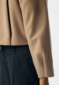 Beige fleece top med en afslappet pasform, der har en side-slids. Sæt sammen med teksturerede mørkeblå bukser med baglommer og subtile linjer.