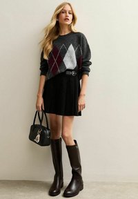 Maglione grigio con motivo a diamante multicolore, gonna plissettata nera, stivali marroni fino al ginocchio e una borsa nera con un charm decorativo.