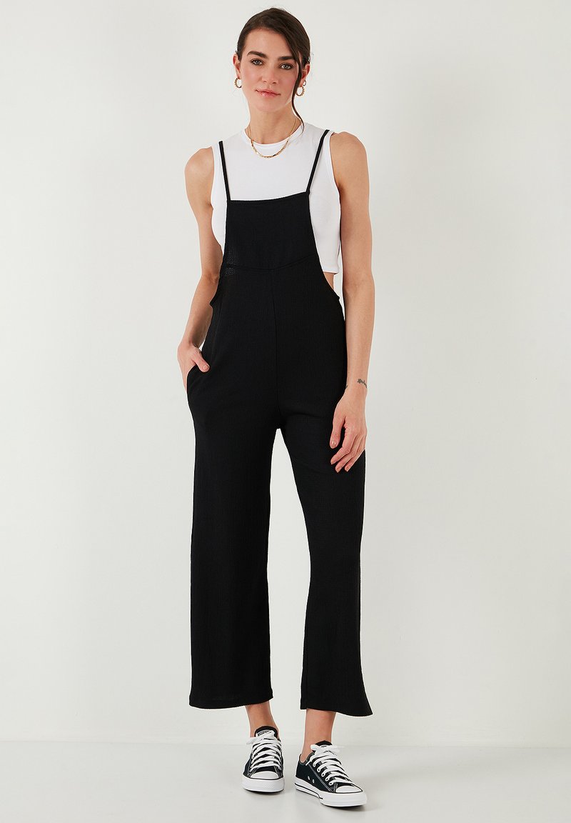Zwarte overall jumpsuit met brede benen en verstelbare bandjes, gecombineerd met een aansluitend wit tanktop en zwart-witte sneakers.
