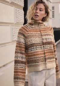 Pull court marron et crème avec un col montant, présentant des motifs géométriques complexes et un tricot texturé. Manches longues avec poignets côtelés.