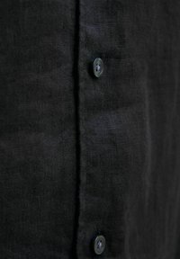 Camisa de lino negra con una superficie texturada y una fila de tres botones. Presenta un diseño limpio y minimalista, con detalles de costura visibles.