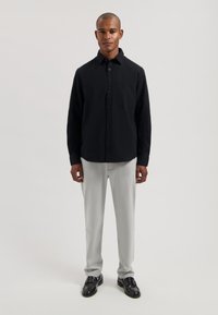 Zwart overhemd met knopen en een gestructureerd patroon, gecombineerd met lichtgrijze broek en zwarte schoenen. Het overhemd heeft een borstzak.