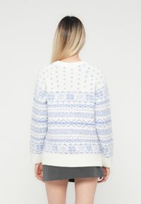 Γυναίκα με μακριά ομπρέ μαλλιά, φορώντας λευκό πουλόβερ με ανοιχτό μπλε ανάγλυφο σχέδιο και κο�ντή σκούρα γκρι φούστα, γυρισμένη μακριά.