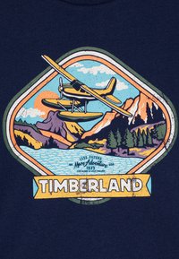 Tamsiai mėlyna marškinė su seaplane grafika virš kalnų ir vandens, su spalvingu saulėtekiu, medžiais ir tekstu "Timberland" apačioje.