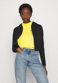 Sudadera negra corta con cremallera frontal, combinada con un cuello alto amarillo brillante con un diseño gráfico. Unos vaqueros de tiro alto de color azul claro completan el look.