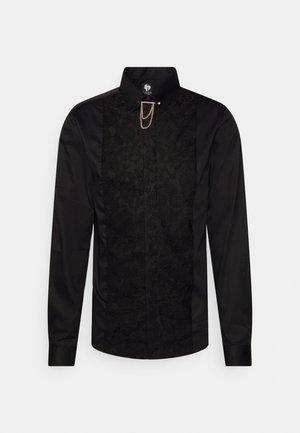 Chemise noire à manches longues avec un panneau avant en dentelle texturée, un col montant et un détail en chaîne dorée fixé au col.