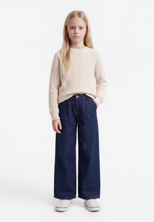 Fată tânără cu păr lung și blond, purtând un pulover beige deschis, pantaloni largi albastru închis și pantofi albi, stând cu o mână în buzunar.