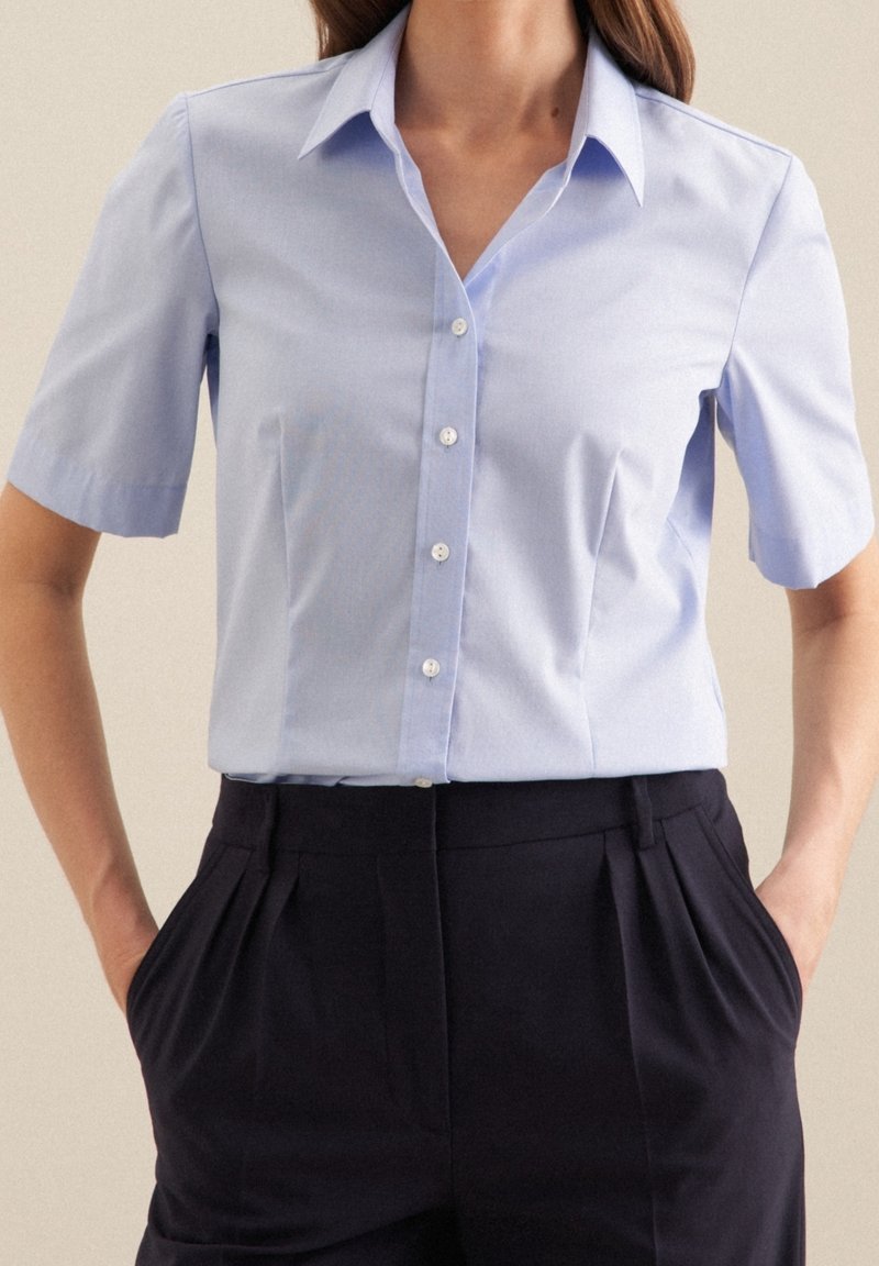 Ljusblå kortärmad skjorta med knäppning framtill, krage och mjuk textur, i kombination med mörka veckade byxor.