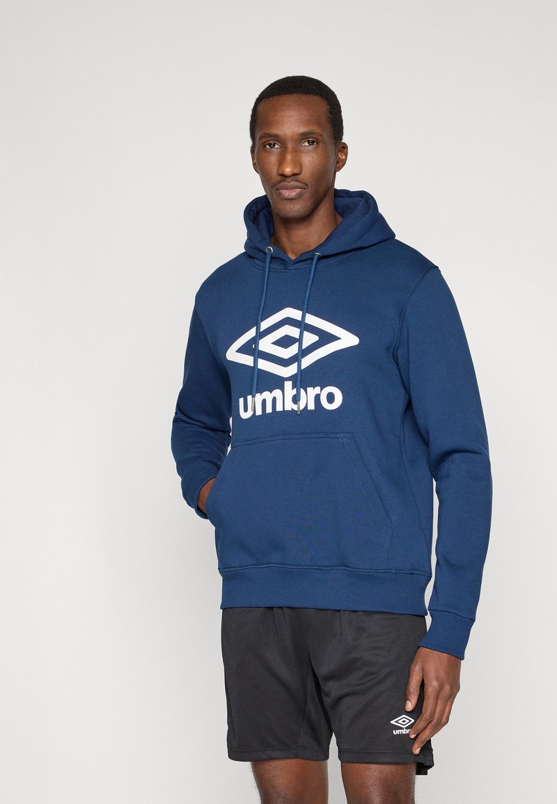 Homme portant un sweat à capuche bleu marine Umbro avec logo et un short noir Umbro, debout devant un fond clair uni avec une main dans la poche.