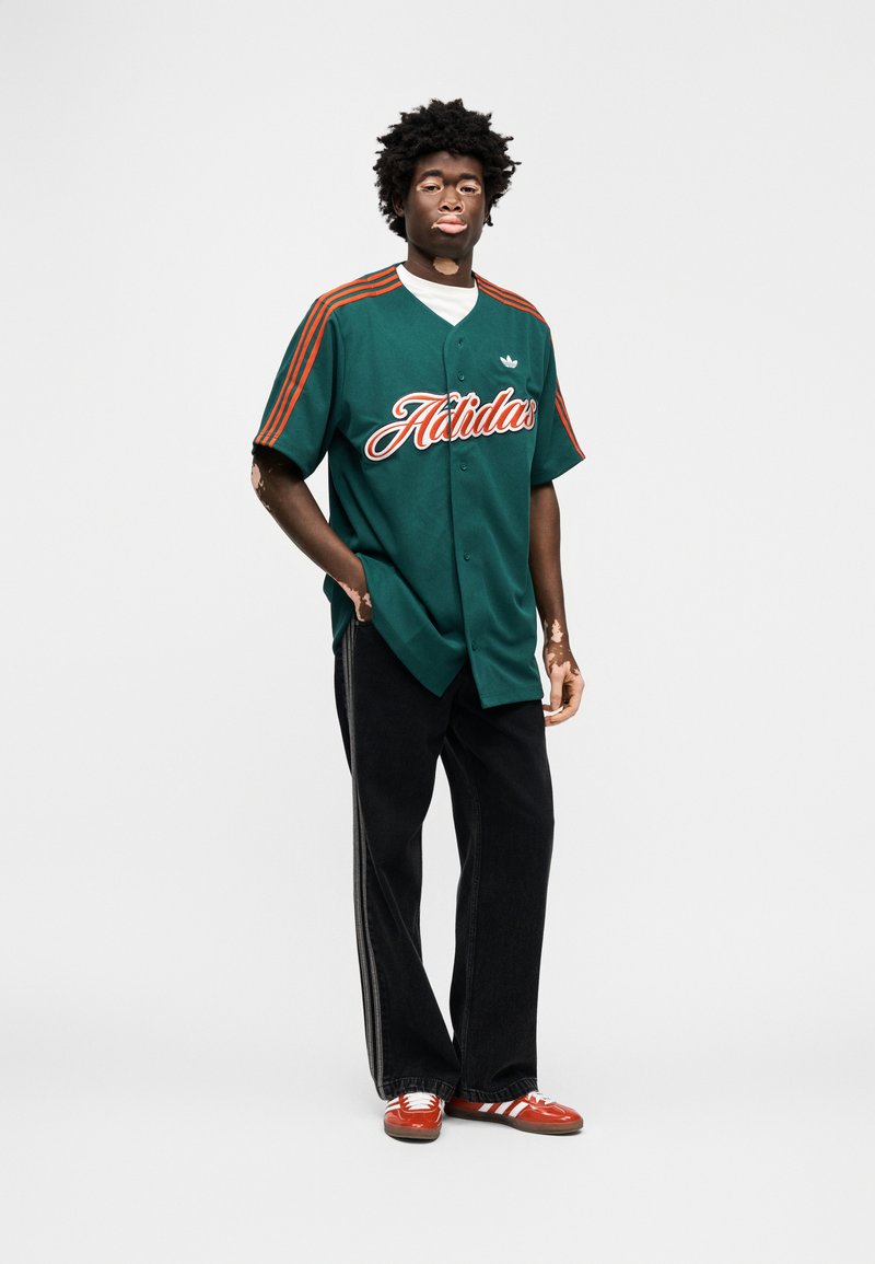 Maillot vert boutonné avec inscription "Holiday" orange et trois bandes orange sur les manches, associé à un pantalon noir et des baskets rouges avec des bandes blanches.