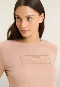 T-shirt rosa chiaro realizzato in un morbido tessuto, con una toppa rettangolare con testo e dettagli in strass sul davanti.
