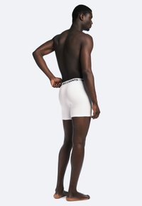 Un short en coton blanc avec une ceinture noire portant le logo "ALLSAINTS". Le modèle est présenté de profil, pieds nus, dans une pose neutre.