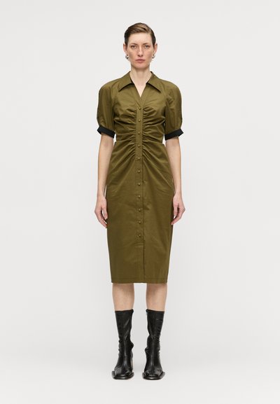 Robe vert olive à manches courtes bouffantes et côtés froncés, associée à des bottes noires montant jusqu'aux genoux. Tissu lisse et texturé.