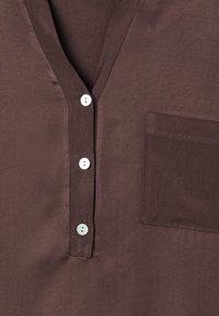 Blusa in raso marrone con scollatura a V, cinque bottoni bianchi e taschino sul petto. Il tessuto ha una texture liscia e una morbida lucentezza.