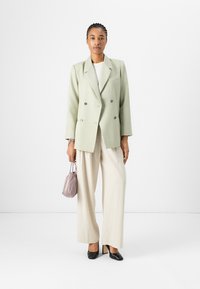 Dvoudílný světle zelený blazer, krémové volné kalhoty, černé lodičky, drží malou levandulovou tašku; hladká tkanina, strukturovaný design.