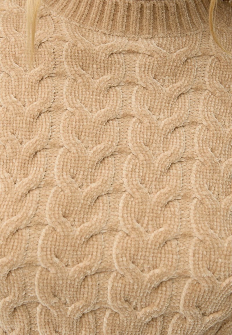Primo piano di un maglione beige a lavorazione chunky con spesse trame a treccia e collo a dolce vita a coste.