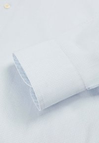 Manga de camisa de vestir azul claro con un fino patrón a cuadros, mostrando el puño con costura prolija y un botón en el cuello.