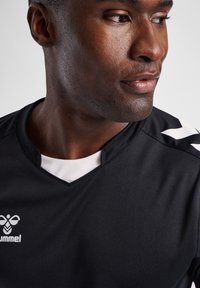Man som bär en svart sporttröja med vitt logotyp och chevronmönster på axeln, tittar åt sidan mot en enfärgad bakgrund.