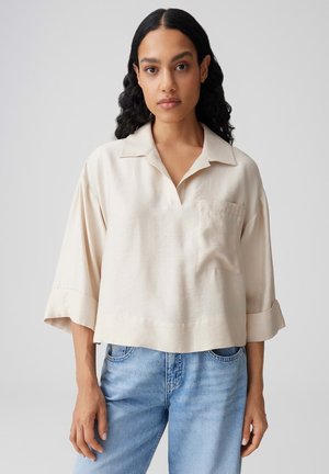 Vrouw met lang krullend zwart haar, gekleed in een loszittend beige overhemd met kraag en brede mouwen en lichtblauwe spijkerbroek, staand tegen een effen achtergrond.