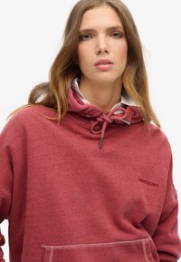 Sudadera con capucha roja hecha de una tela texturizada. Presenta un cierre con cordones, un bolsillo frontal y un logo bordado en el pecho.
