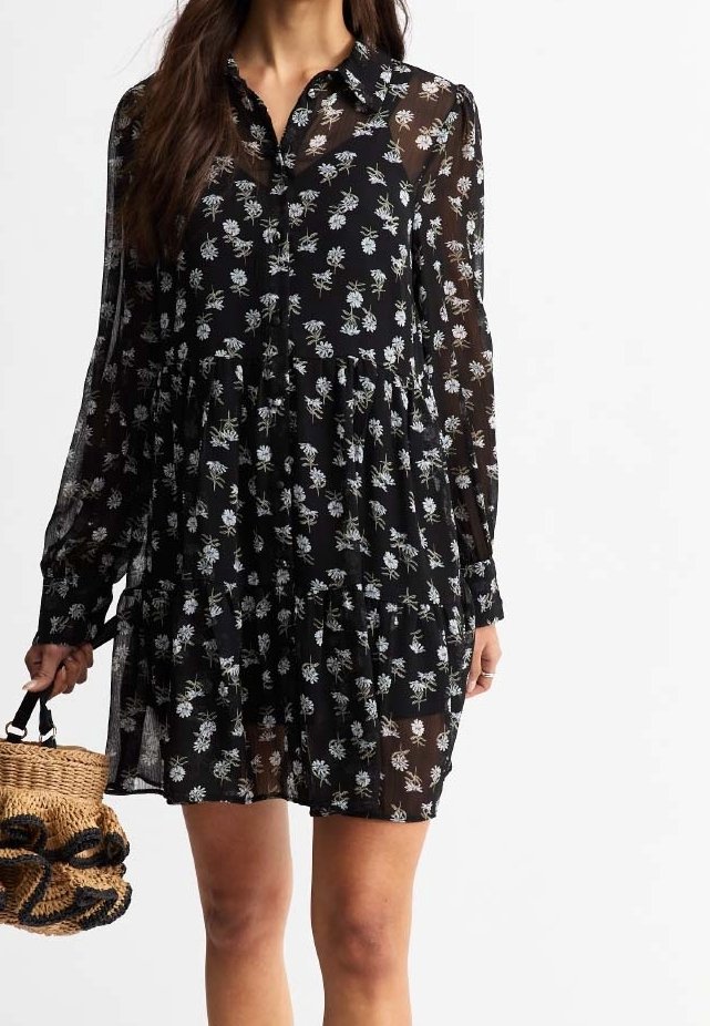 Abito nero trasparente con stampa floreale, maniche lunghe, colletto con bottoni, vita arricciata e gonna a balze. Abbinato a una borsa intrecciata.