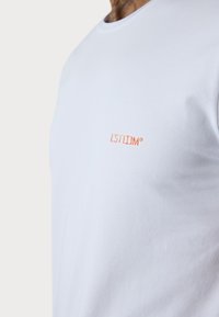 Weißes T-Shirt aus weichem Material mit rundem Ausschnitt und kleinem, orangefarbenem Logo, das auf der Brust gestickt ist. Minimalistisches Design.