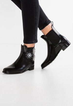 Sorte blanke ankelstøvler med blomsterdekorationer båret med sorte tætsiddende bukser på en hvid gulv.