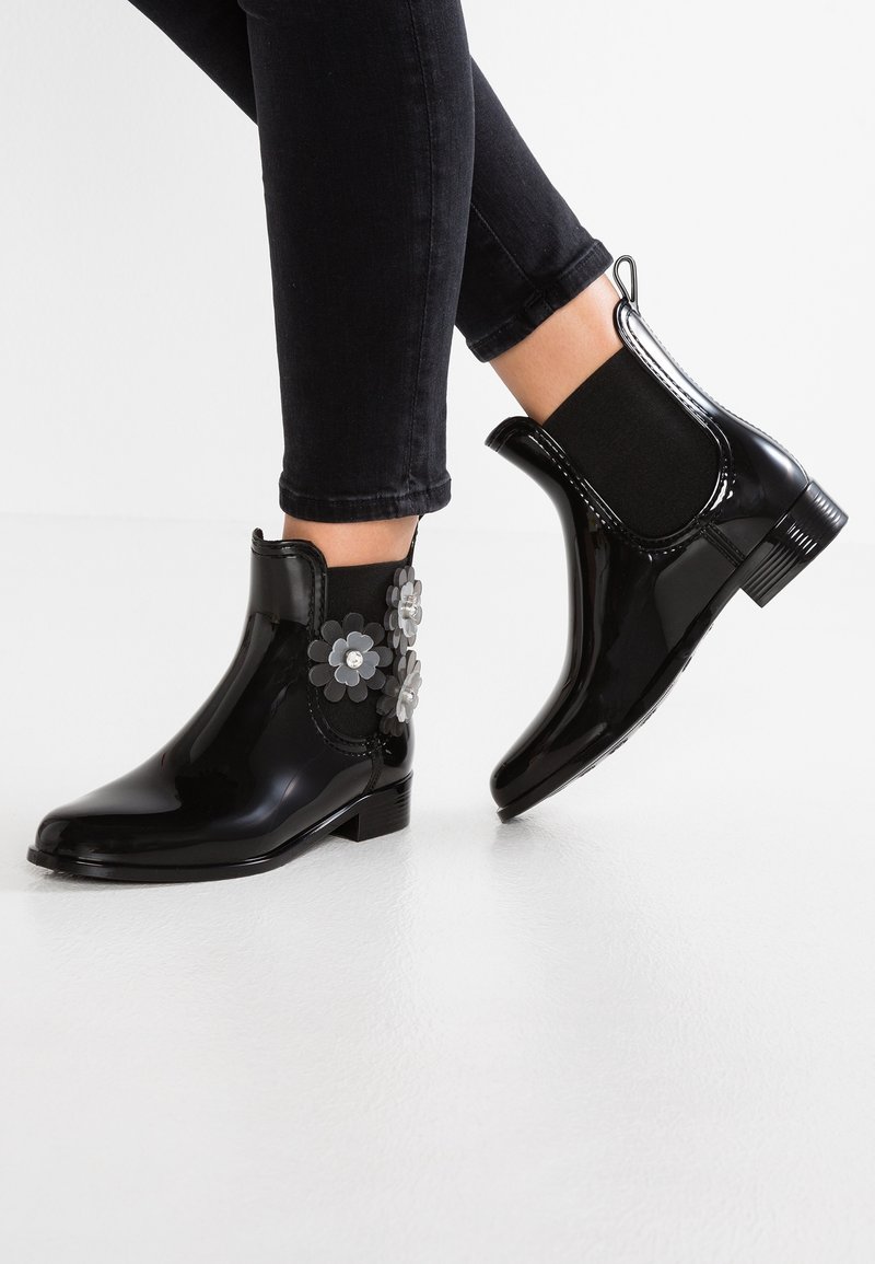 Bottines noires brillantes avec décorations florales portées avec un pantalon noir ajusté sur un sol blanc.