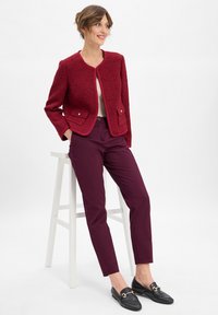 Giacca in tweed rosso con scollo rotondo, due tasche frontali e bottoni argentati, abbinata a pantaloni bordeaux e mocassini neri.