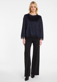 Marineblauer Fransenpullover mit Rundhalsausschnitt und langen Ärmeln, kombiniert mit hochgeschnittenen schwarzen Hosen, die feine Glitzereffekte aufweisen.