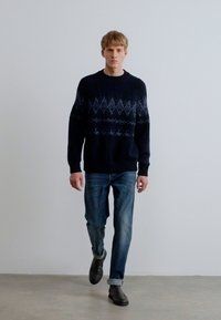 Tengerészkék kötött pulóver, amelynek felső részén fair isle mintázat található, világoskék farmernadrággal és fekete cipővel párosítva.