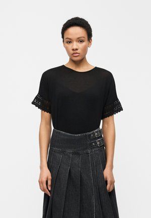 Kvinde med kort krøllet hår iført en sort top med blonder på ærmerne og en mørk plisseret højhalset nederdel med spænde detaljer.