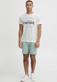 Hvit bomull t-skjorte med grafikk "BLEND DENIM ORIGINAL VINTAGE EST. 1993". Lysegrønne shorts, hvite joggesko, stående posisjon.