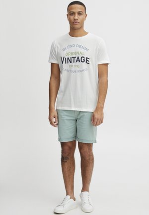 White cotton t-shirt with "BLEND DENIM ORIGINAL VINTAGE EST. 1993" graphic. Light green shorts, white sneakers, standing pose.