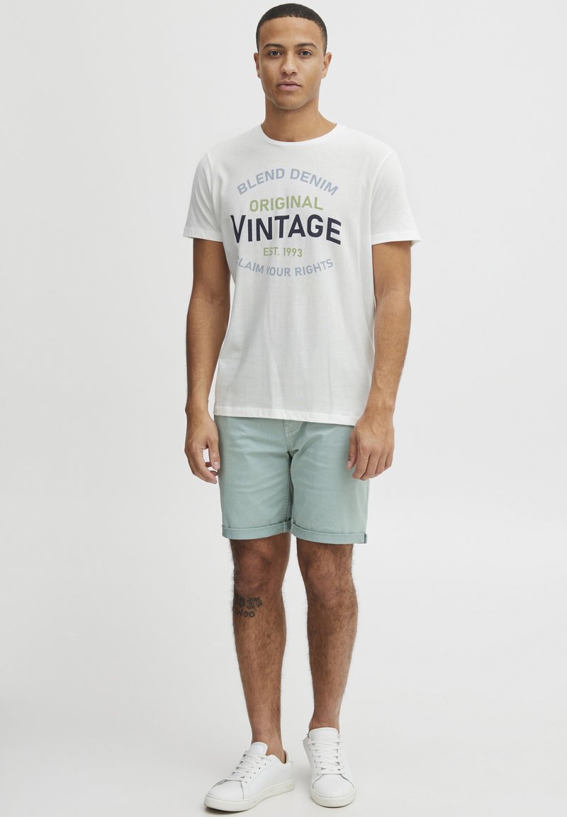 Valkoinen puuvillapaita, jossa on "BLEND DENIM ORIGINAL VINTAGE EST. 1993" -grafiikka. Vaaleanvihreät shortsit, valkoiset tennarit, seisovassa asennossa.