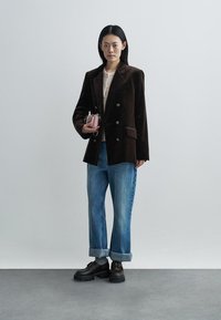 Blazer castanho de bombazina com botões duplos, combinado com jeans azul claro com bainhas viradas e sapatos pretos robustos. Segurando uma mala rosa.