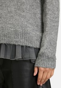 Pull en tricot gris avec ourlet et poignets côtelés, superposé à un tissu noir transparent à volants. La texture est douce et légèrement duveteuse.