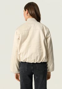 Beige oversized bomberjack met een geribbelde kraag, elastische zoom en geborduurde textuur, gecombineerd met donkergrijze high-waisted jeans.