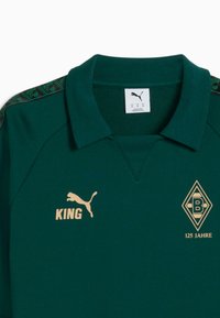 Tricou polo verde din bumbac, cu guler. Accente aurii care includ textul 'KING' și un logo cu '125 JAHRE'. Detalii texturate pe umeri.