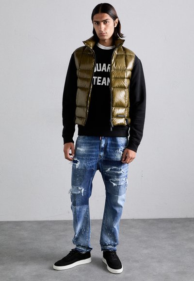 Puffy olive green vest over a black sweatshirt, worn with distressed blue jeans. Black sneakers complete the outfit. Simple background.
