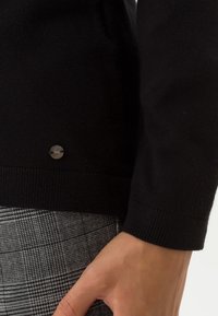 Černý svetr s vroubkovaným lemem a kovovým detailem loga na boku, v kombinaci se šedými kockovanými kalhotami s texturovaným vzorem.