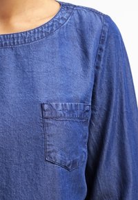 Chemise en jean bleu avec encolure arrondie, manches longues et poche poitrine au design structuré en tissu texturé.