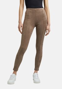 Vrouw in high-waisted, bruine suedine leggings met naaddetails, gecombineerd met witte sneakers en een zwarte mouwloze top.