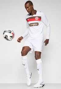 Hvit fotballdrakt med røde og brune detaljer, lange ermer, med et logo og sponsortekst. Korte hvite shorts, hvite sokker og svarte fotballsko.