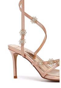 Sandali con tacco alto color oro rosato, dotati di cinturini metallici e decorazioni con strass. Design con punta aperta, tacco sottile, texture liscia.