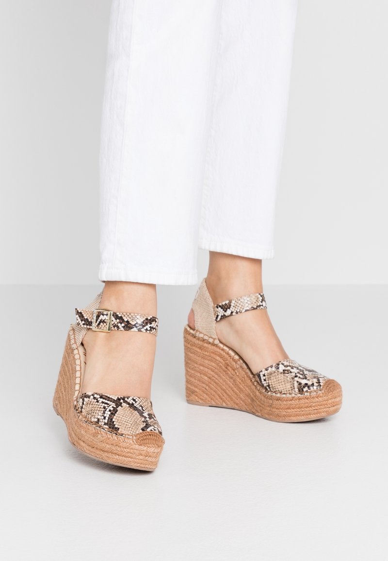 Sandales compensées avec brides à imprimé serpent et talons compensés en jute portées avec un pantalon blanc court sur un sol lisse.