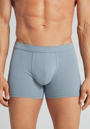 Mandekrop iført lyseblå tætsiddende boxershorts mod en ensfarvet baggrund.
