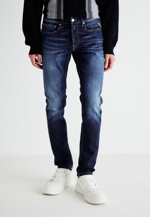 Personne portant un jean slim bleu foncé et des baskets blanches, debout devant un fond clair uni.
